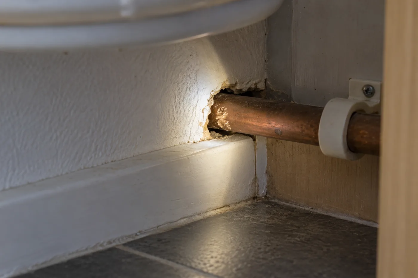 Gap around a copper pipe beneath a kitchen cabinet highlighted by a torch
